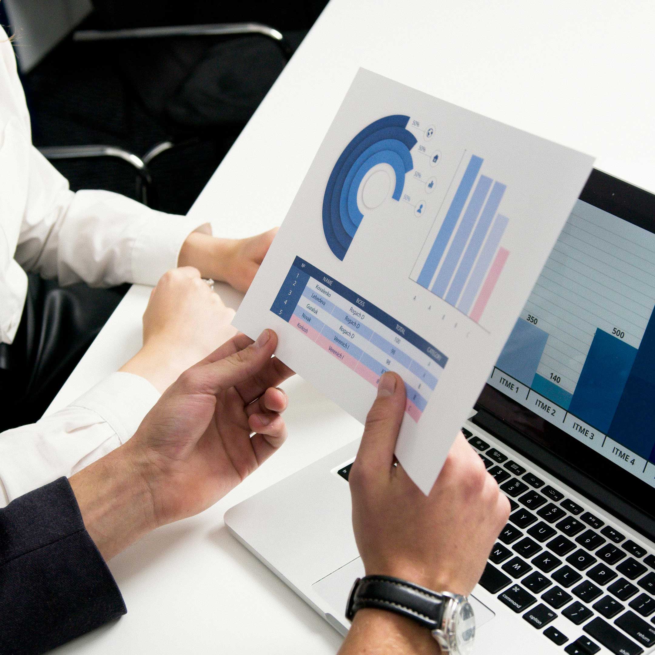 Image of two people sitting at a desk reviewing financial data printed on a piece of paper with a laptop within reach